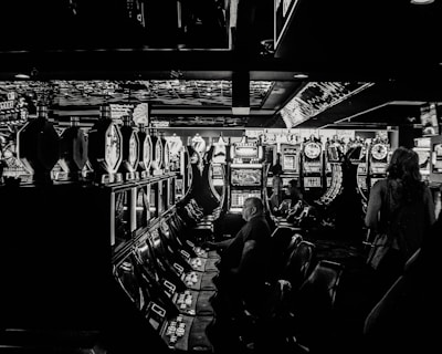A dimly lit casino filled with rows of slot machines, some occupied by people. The machines' lights contrast with the dark surroundings, creating a vibrant yet subdued atmosphere.