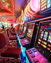 A brightly lit casino interior featuring a row of colorful slot machines. The neon lights create a vibrant atmosphere, with patterned carpeting adding to the decor. Stools are aligned neatly in front of each machine.