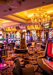 A vibrant casino interior filled with rows of colorful slot machines, each illuminated with bright lights and displays. The ceiling is adorned with elegant chandeliers and golden drapes, creating a luxurious atmosphere. The patterned carpet adds to the elaborate design of the space.
