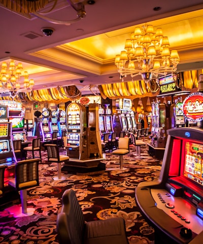 A vibrant casino interior filled with rows of colorful slot machines, each illuminated with bright lights and displays. The ceiling is adorned with elegant chandeliers and golden drapes, creating a luxurious atmosphere. The patterned carpet adds to the elaborate design of the space.