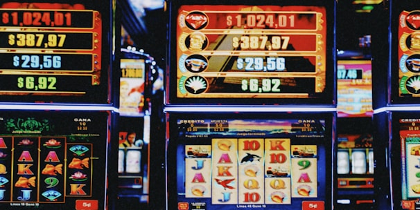 A row of colorful slot machines lit with bright neon lights, showcasing various graphical elements on the screens, indicating different games and jackpots. The atmosphere suggests a lively casino setting, bustling with excitement.