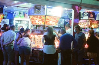 A group of people standing in front of an illuminated amusement arcade, engaging with various gaming machines. The environment is bustling with activity, and colorful lights create a vibrant atmosphere. The ceiling is decorated with large, playful images of dice and other game-related graphics, enhancing the lively mood.